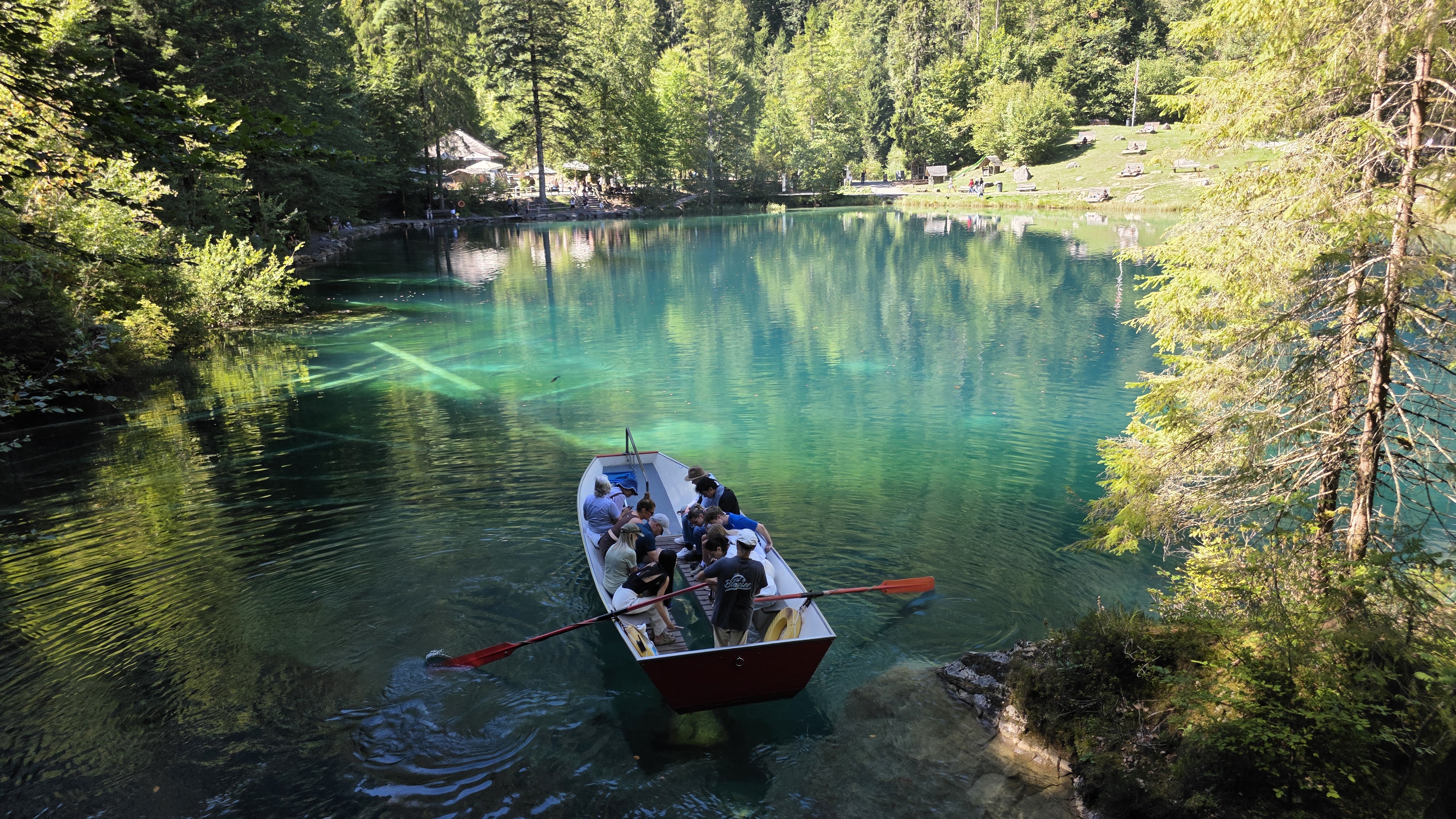 A wooden rowing boat with visitors crossing the turquoise waters of Blausee, with submerged tree trunks visible beneath the surface and forested shoreline in the background.